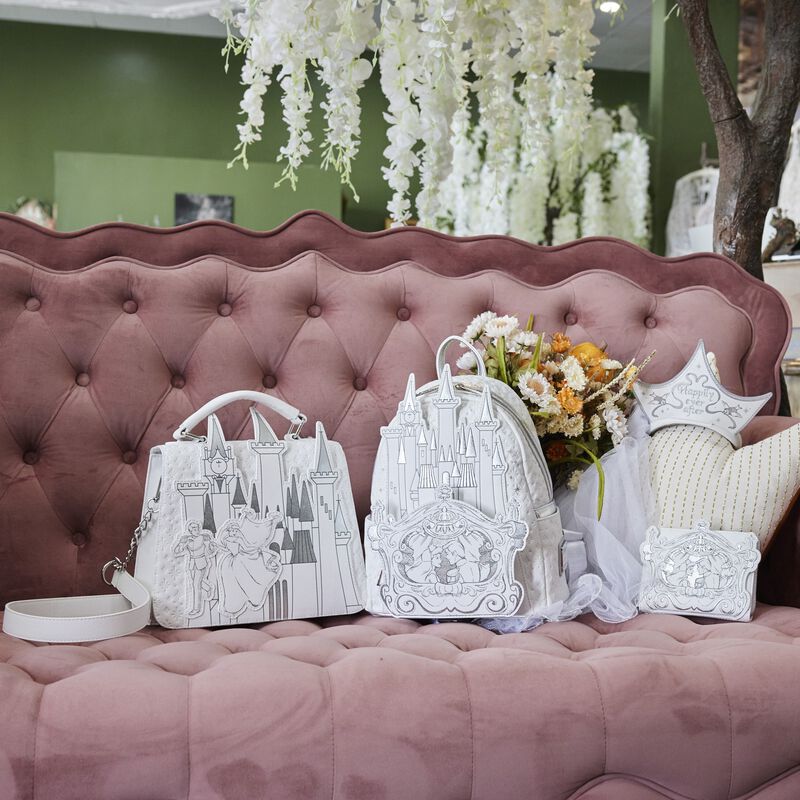Several pieces of the Cinderella Happily Ever After Collection (backpack, crossbody, veil headband, and wallet) sitting together on a pink couch in a bridal shop